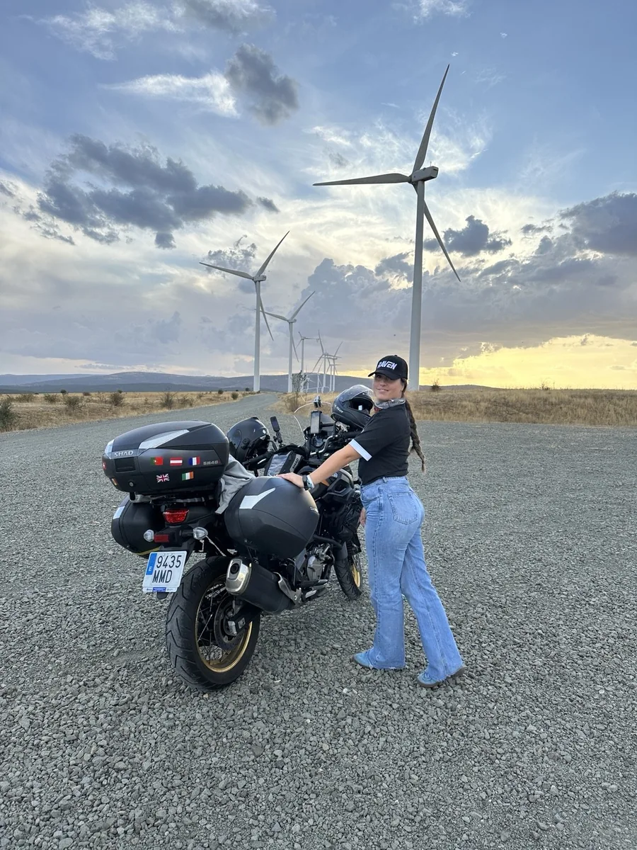 Lara junto a nuestra Suzuki V-Strom en un parque eólico al atardecer